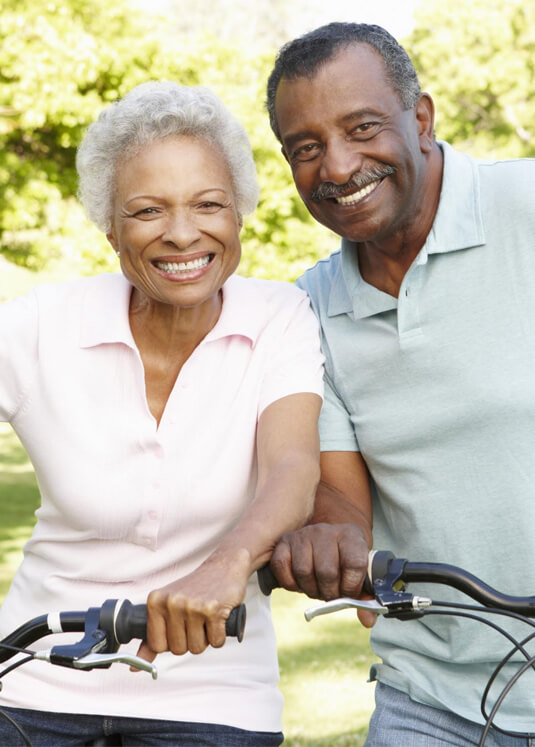 Mature couple smiling while holding bicycles outdoors in sunny park setting.