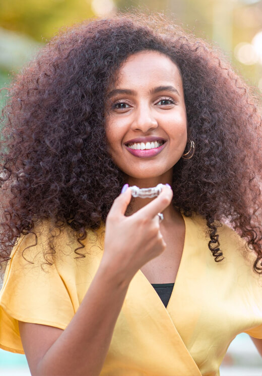 Smiling woman in yellow shirt with hand near chin, outdoors with blurred background.