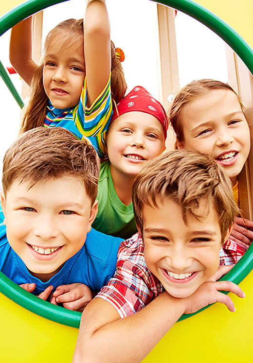 Five smiling children looking down at camera through green playground tunnel opening.