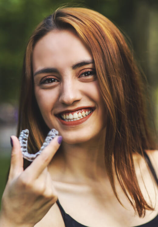 Young woman smiling at camera, holding clear orthodontic aligner, outdoor setting.