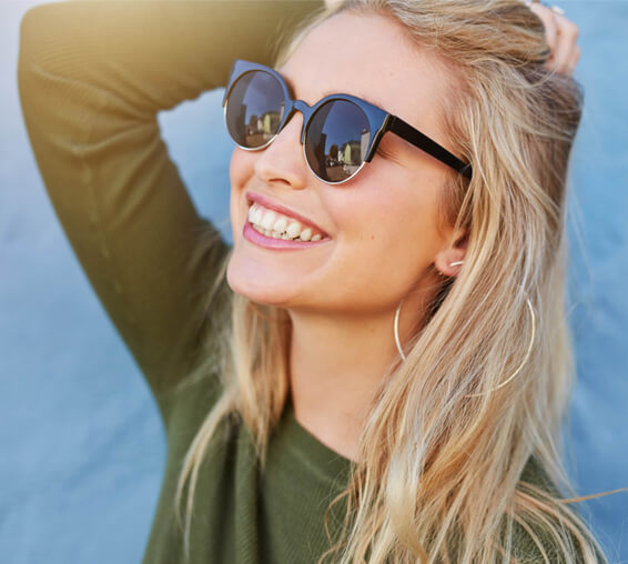 Smiling woman wearing sunglasses and olive green shirt against blue background.