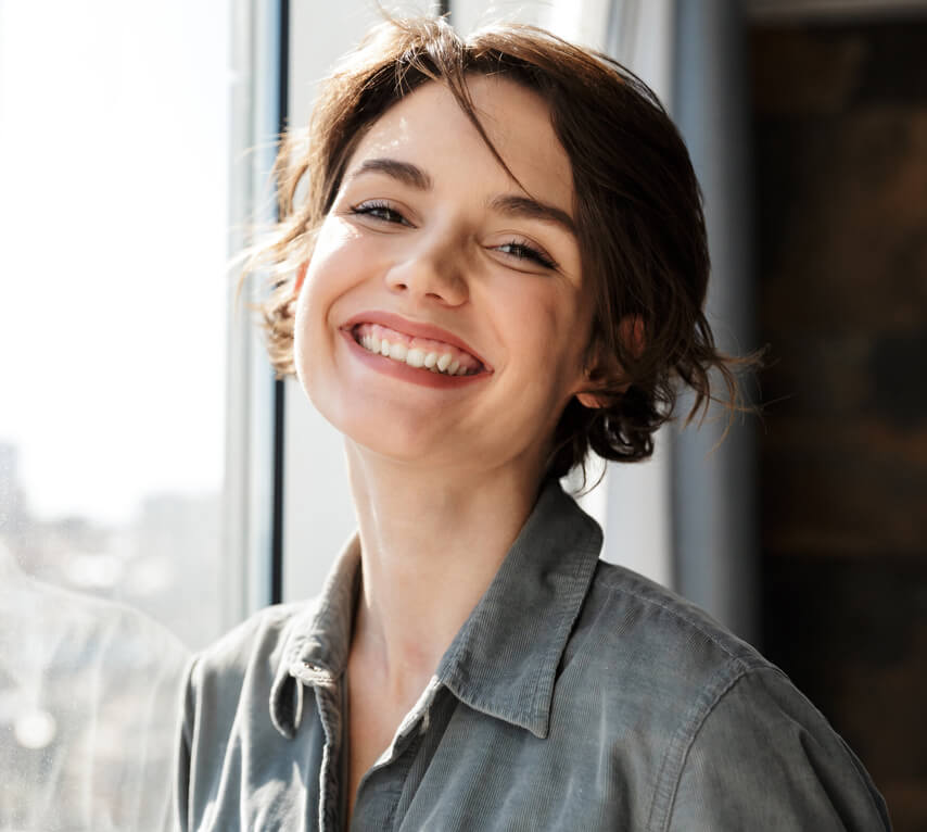 Smiling woman in denim shirt by window in modern dental office setting.