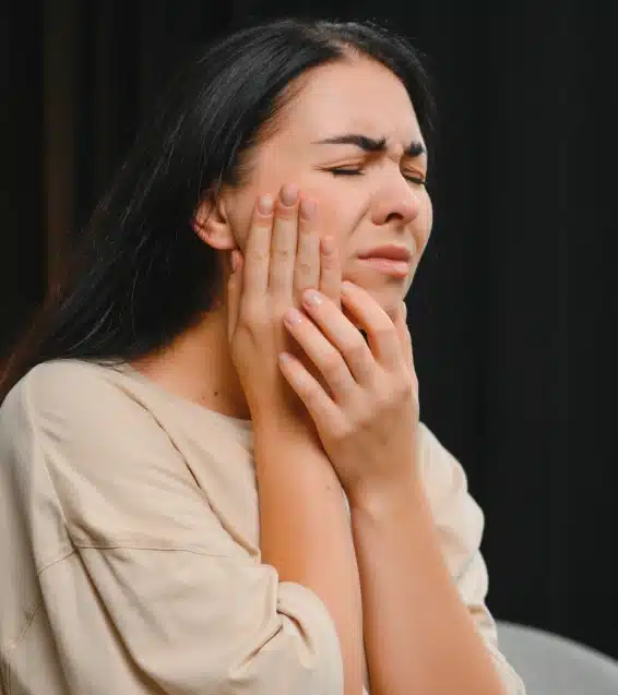 Woman experiencing tooth pain, holding her cheek with both hands against dark background.